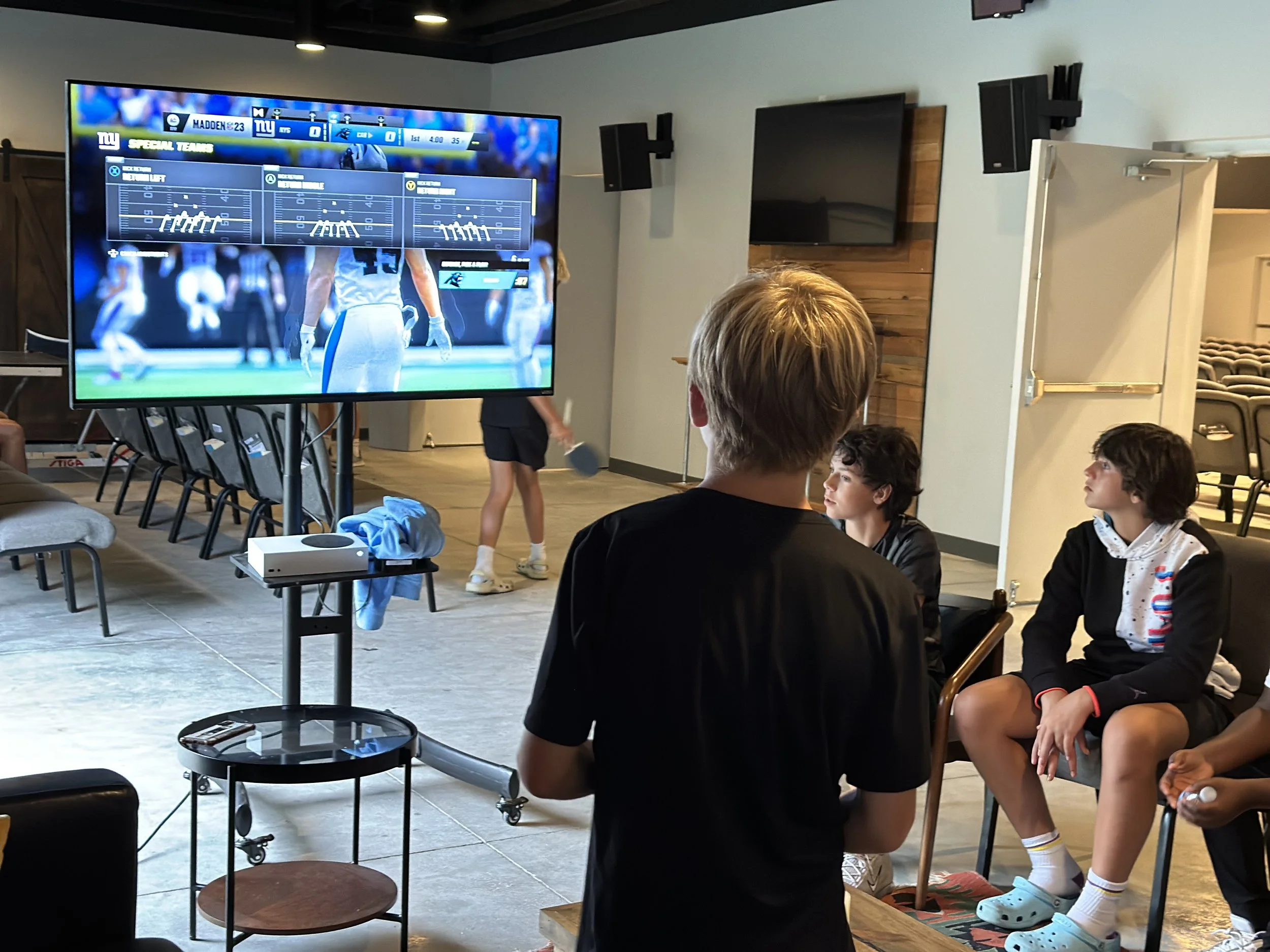 People watching a football game on a large television screen in a lounge or game room, with some individuals seated and others standing nearby.