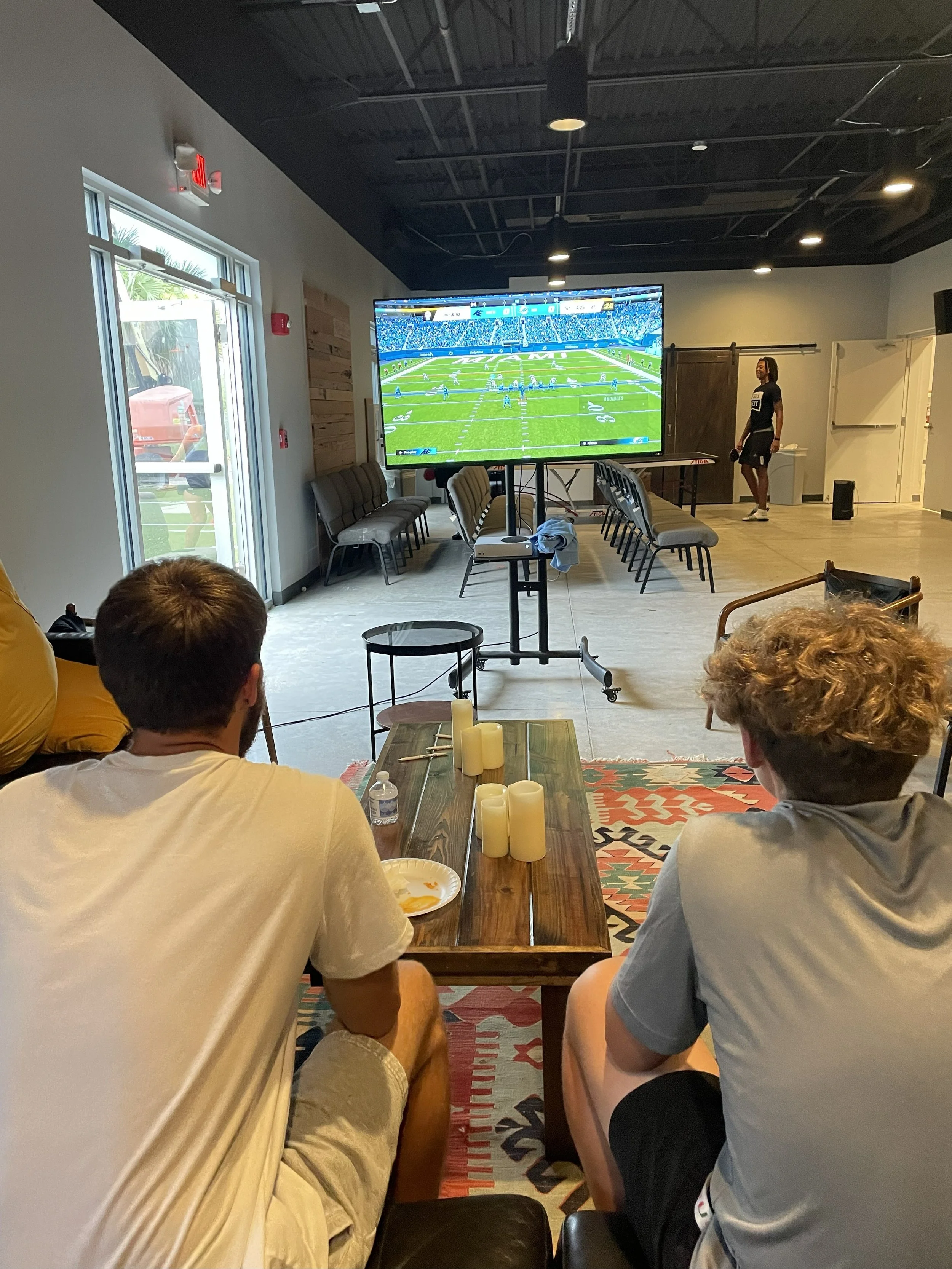 Two people sitting at a wooden table watching a football game on a large flat-screen TV in a room with industrial style ceiling, chairs along the wall, and a person standing near the door.