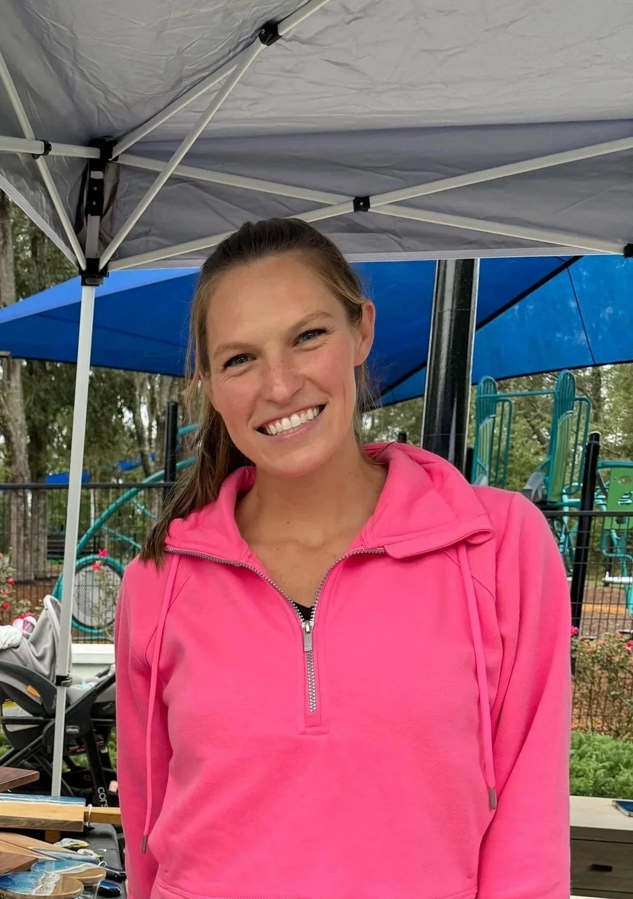 A smiling woman in a pink jacket standing outdoors under a canopy with playground equipment in the background.