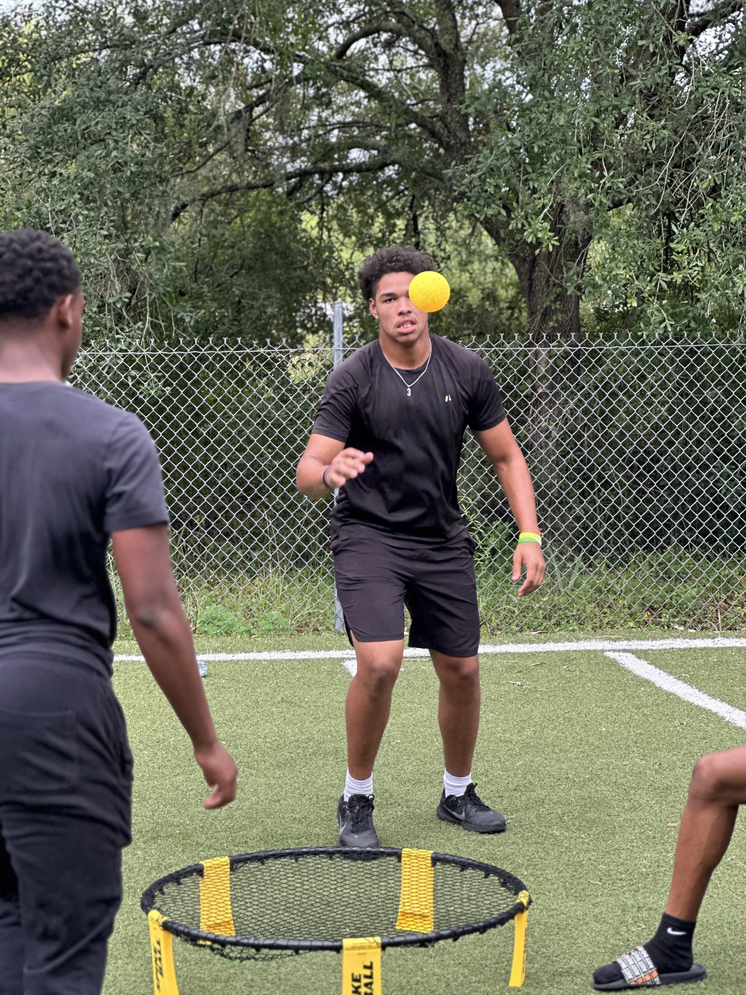 Young man playing cornhole outside, about to toss a yellow bean bag. Two other people are nearby, and a chain-link fence with trees is in the background.