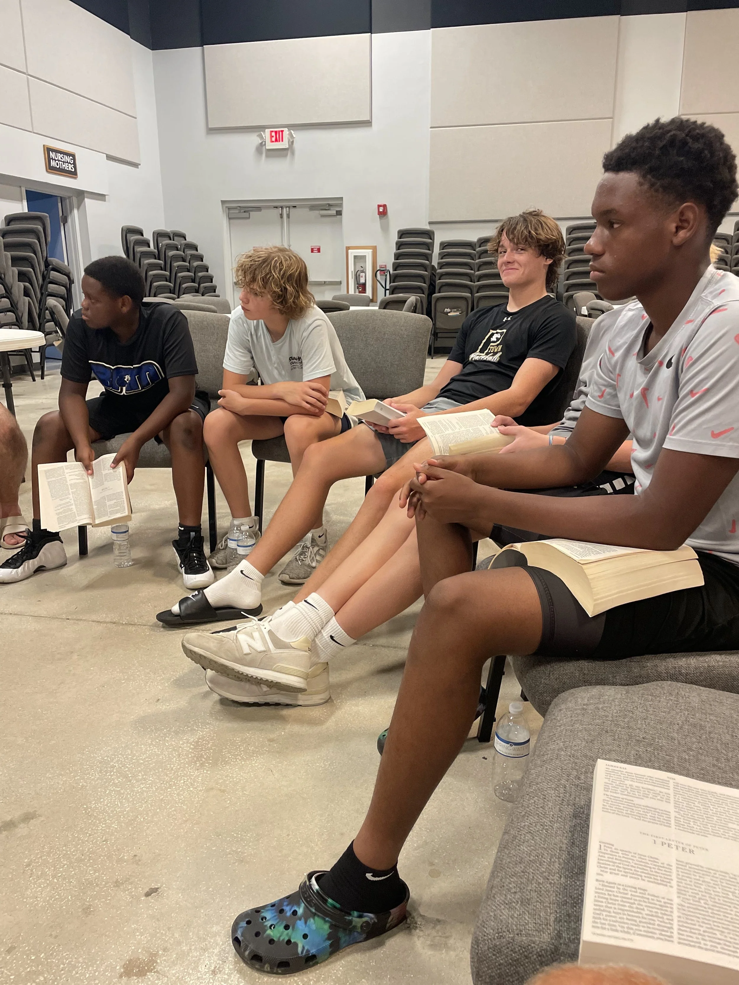 Four teenagers sitting in a circle with books and water bottles in what appears to be a classroom or meeting room, with empty chairs stacked in the background.