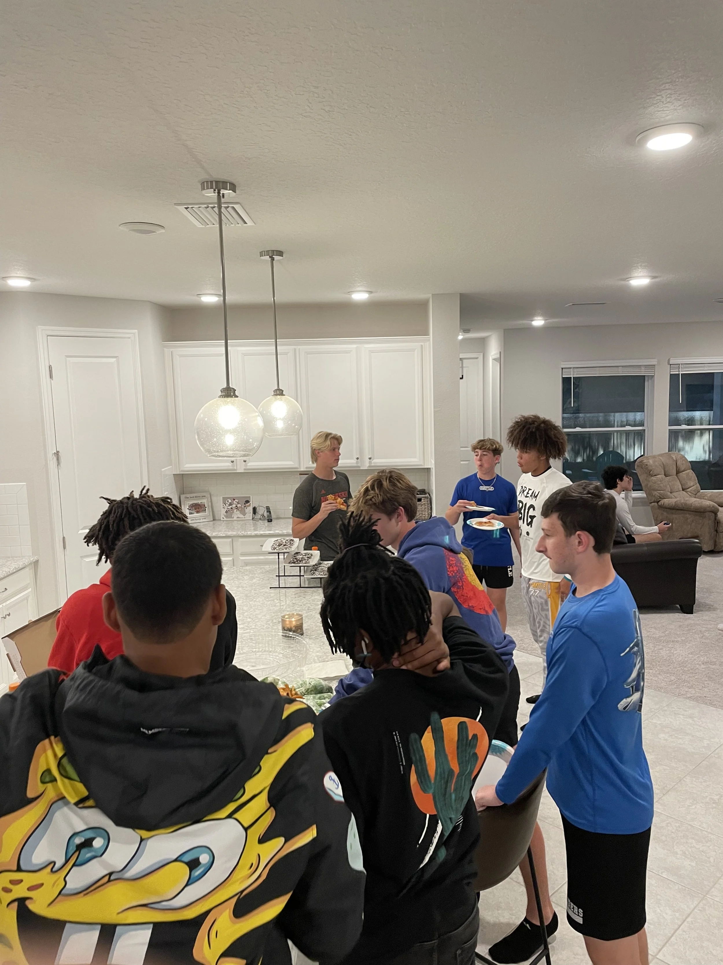 Group of teenagers gathered around a kitchen island, talking and eating in a modern, well-lit home.