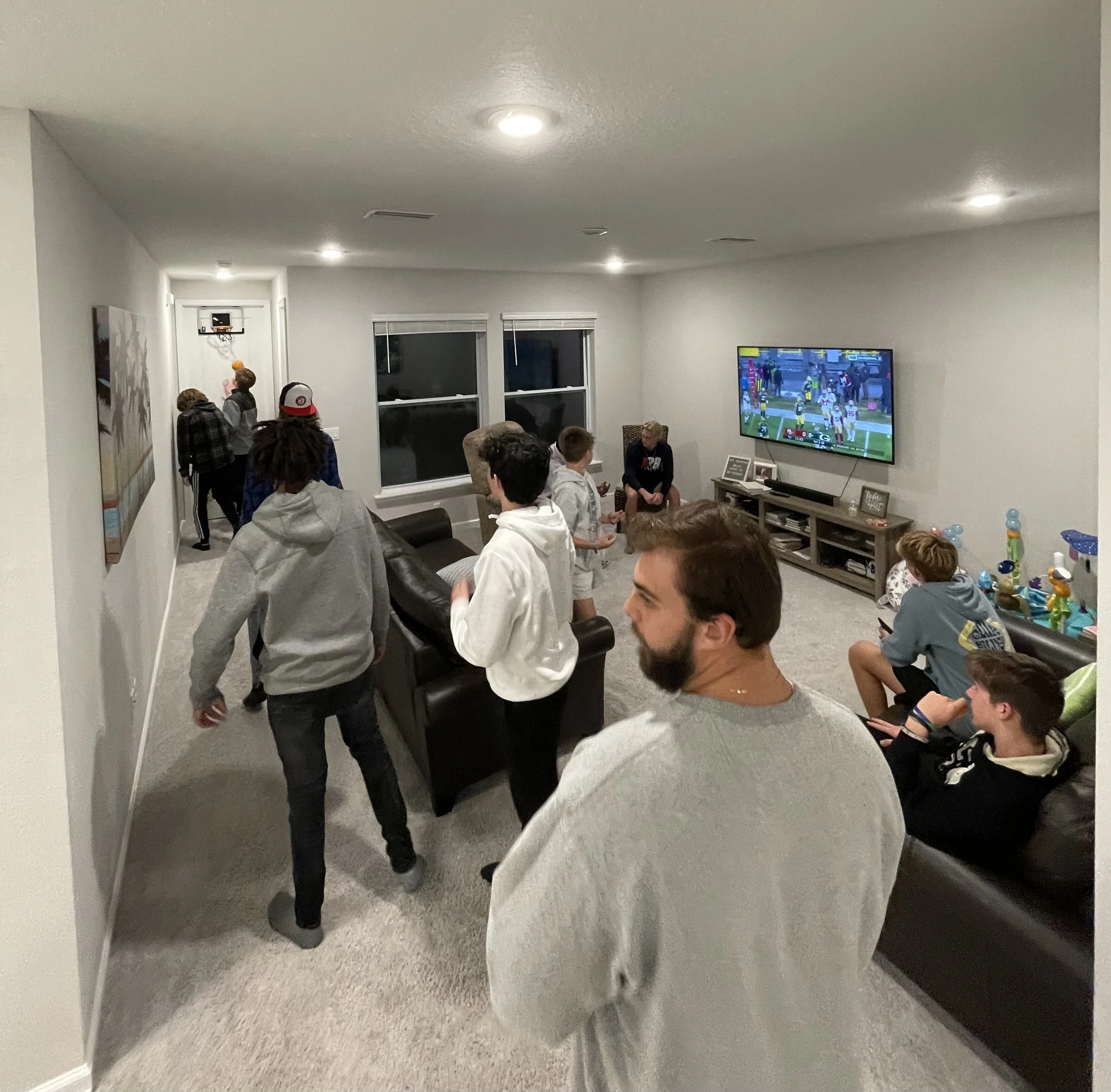 A group of people, mostly teenagers, gathered in a living room watching a football game on TV. Some are sitting on couches, others standing or walking around, with balloons and snacks visible.