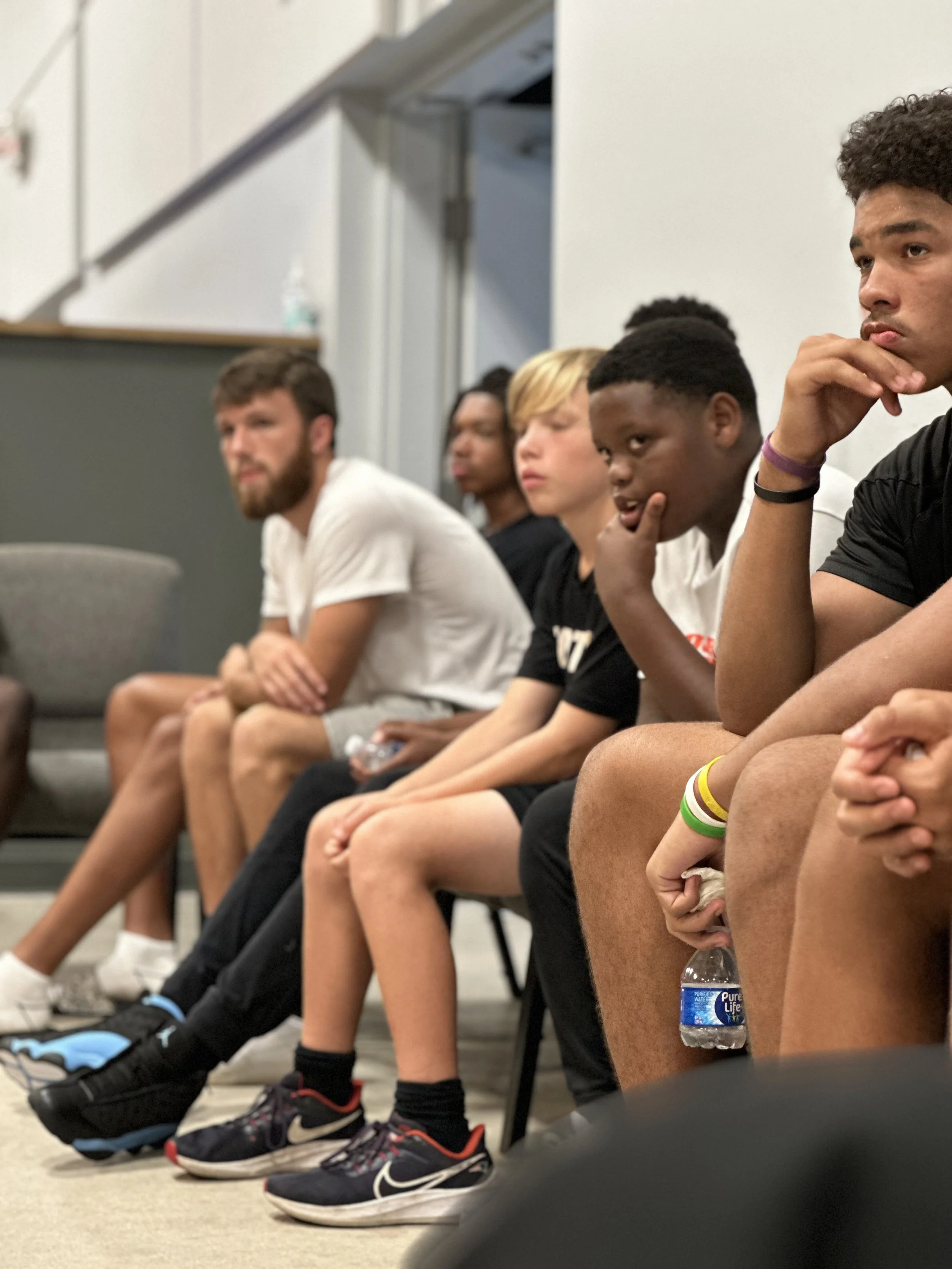 A group of young people sitting in a row on chairs, listening attentively at an indoor event or workshop.