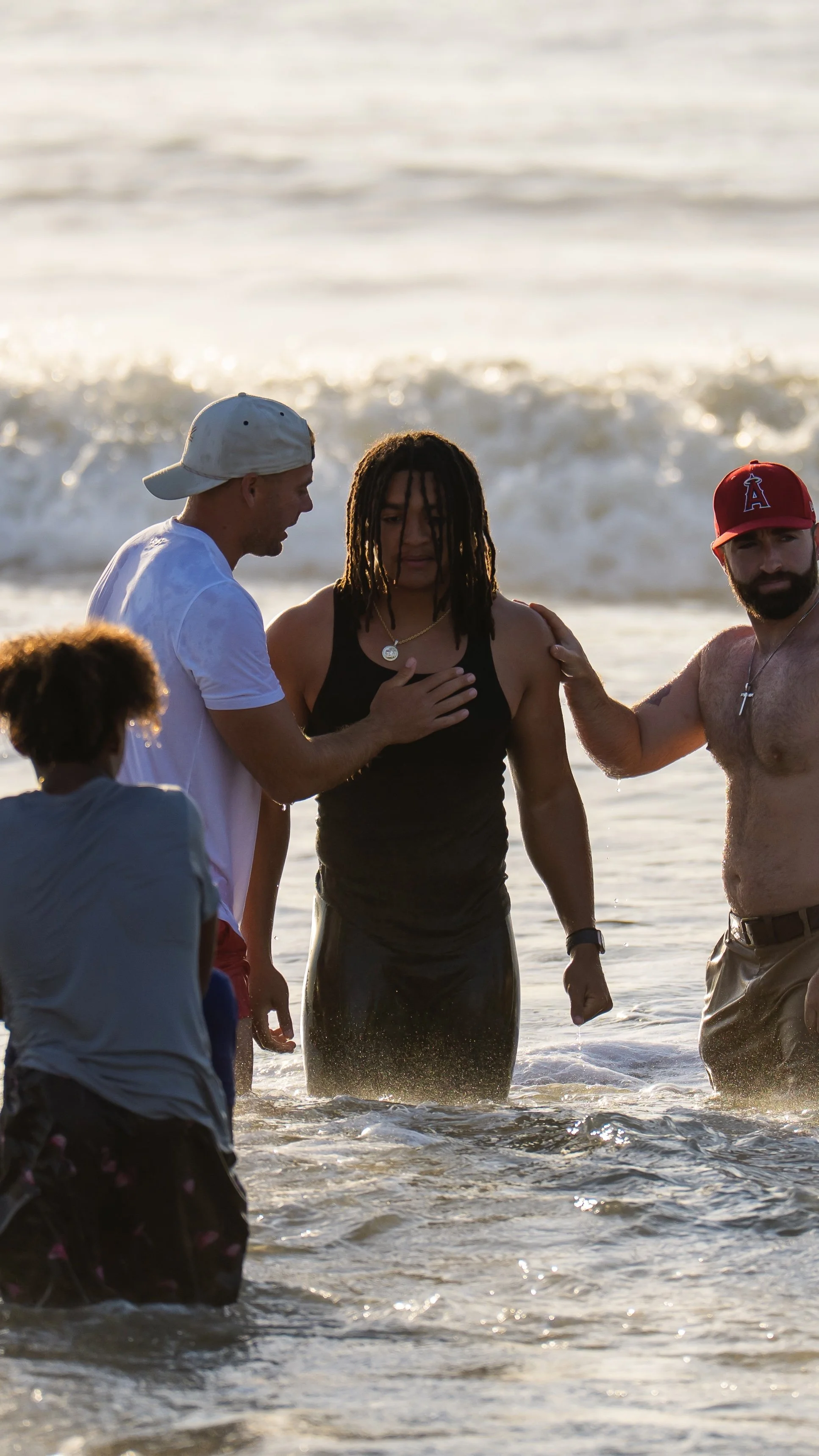A group of people at the beach during sunset, with a woman in a black dress being prayed over or blessed by men, some of whom are shirtless, near the water's edge.