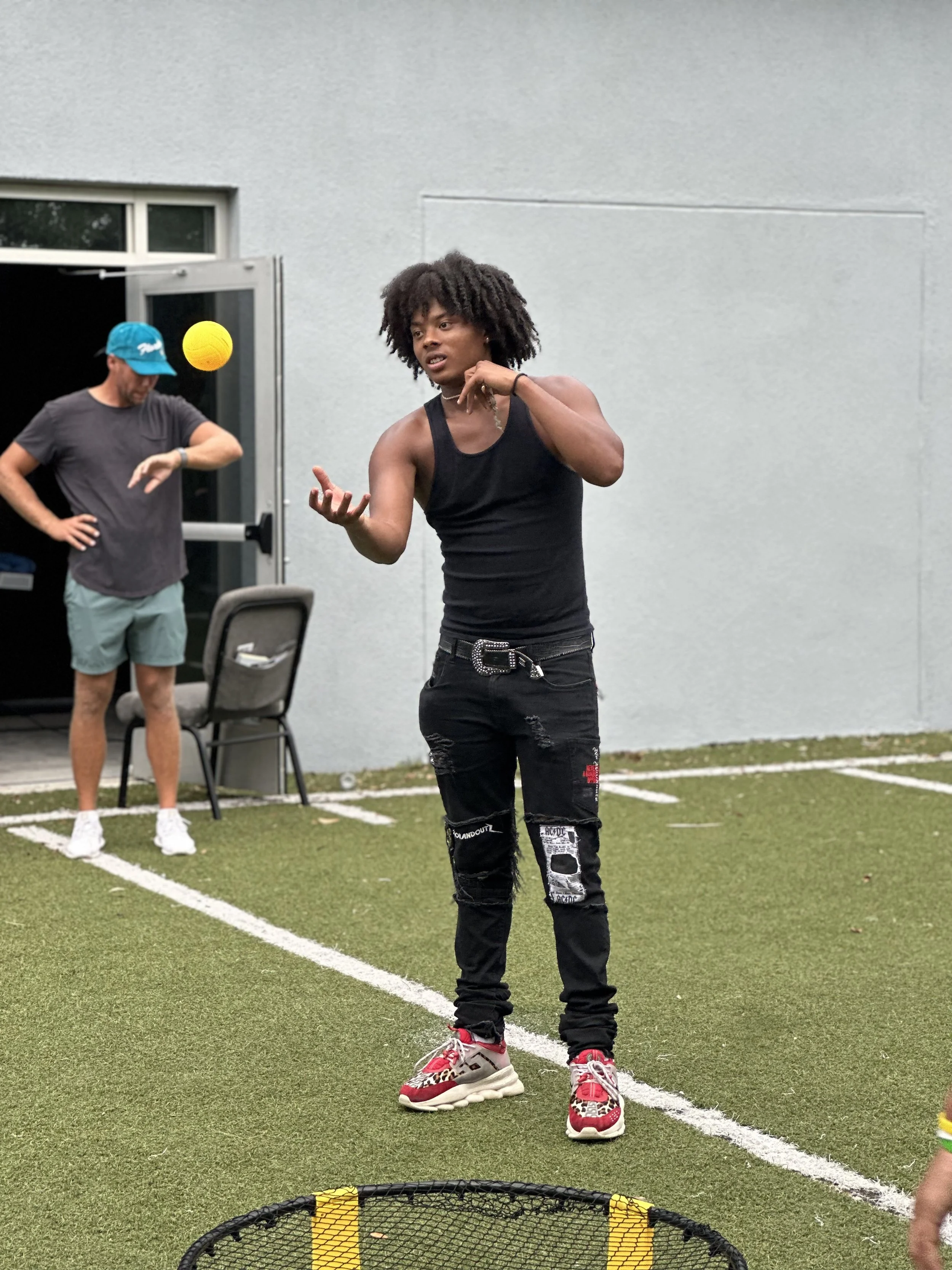 A woman in a black tank top and ripped black jeans is playing a game outside on a grassy area, with a man in a gray t-shirt and teal shorts in the background.