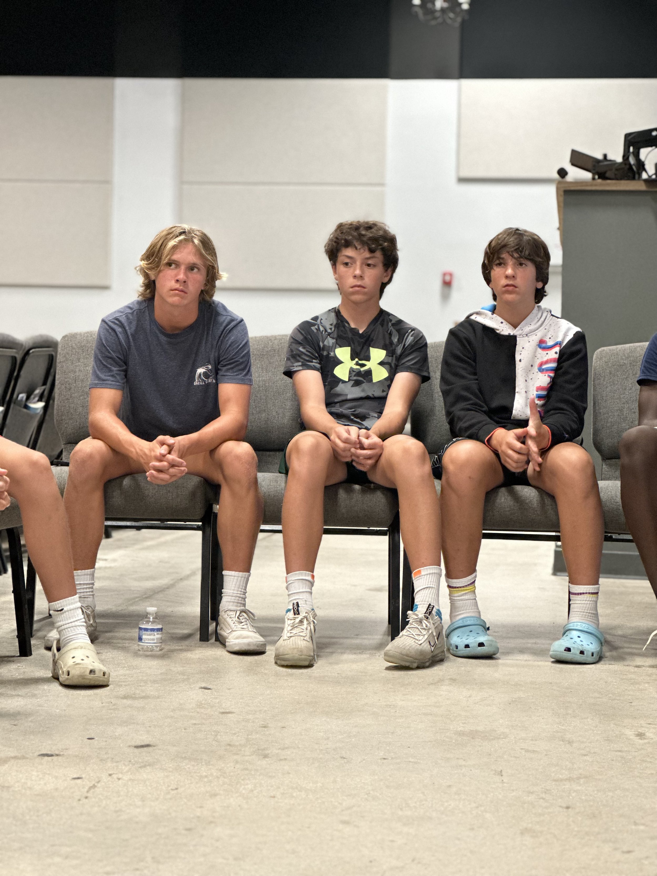 Three boys sitting on a gray couch in an indoor setting, wearing casual shorts and t-shirts, with serious expressions, some with hands clasped or resting on their laps.