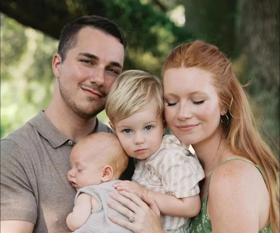 A family portrait of a man, woman, and three children outdoors. The man and woman hold the children, one of whom is a sleeping baby, while the other looks at the camera.