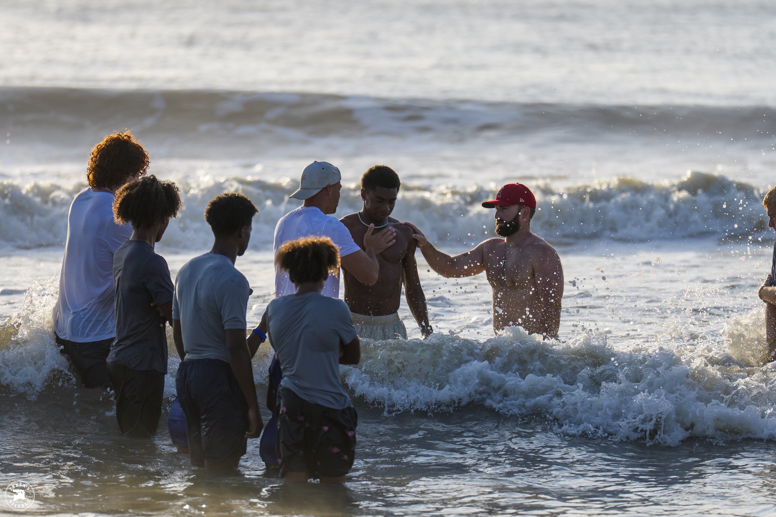 A group of people, including children and adults, standing in the ocean waves, participating in a baptism or religious ceremony with one man submerged and others assisting.