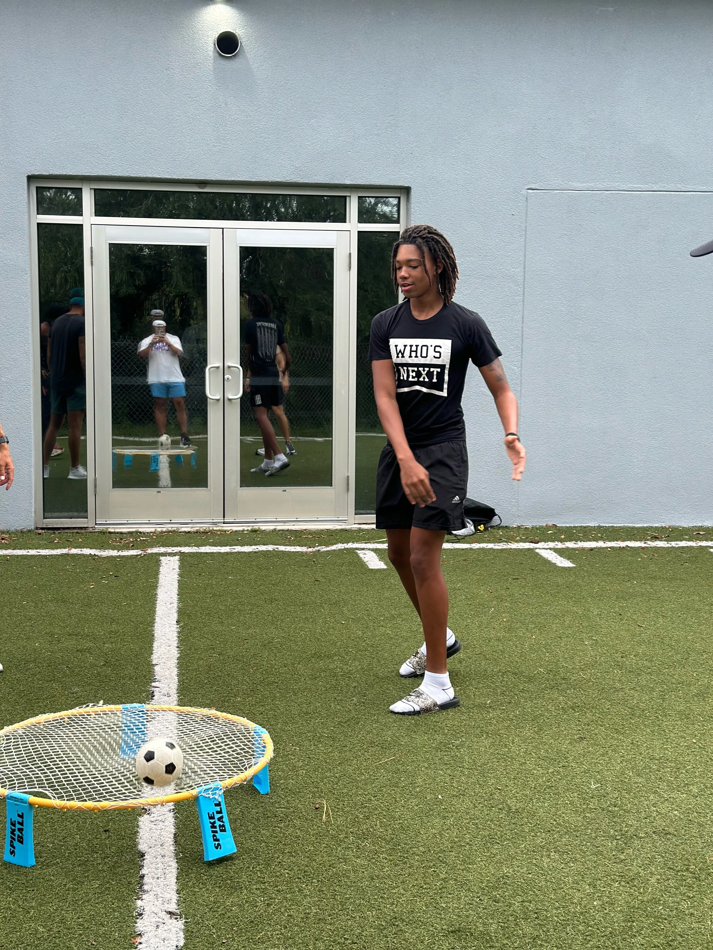 A young man playing spikeball on a grassy outdoor court, with a small soccer ball in mid-air above the net, during daytime.