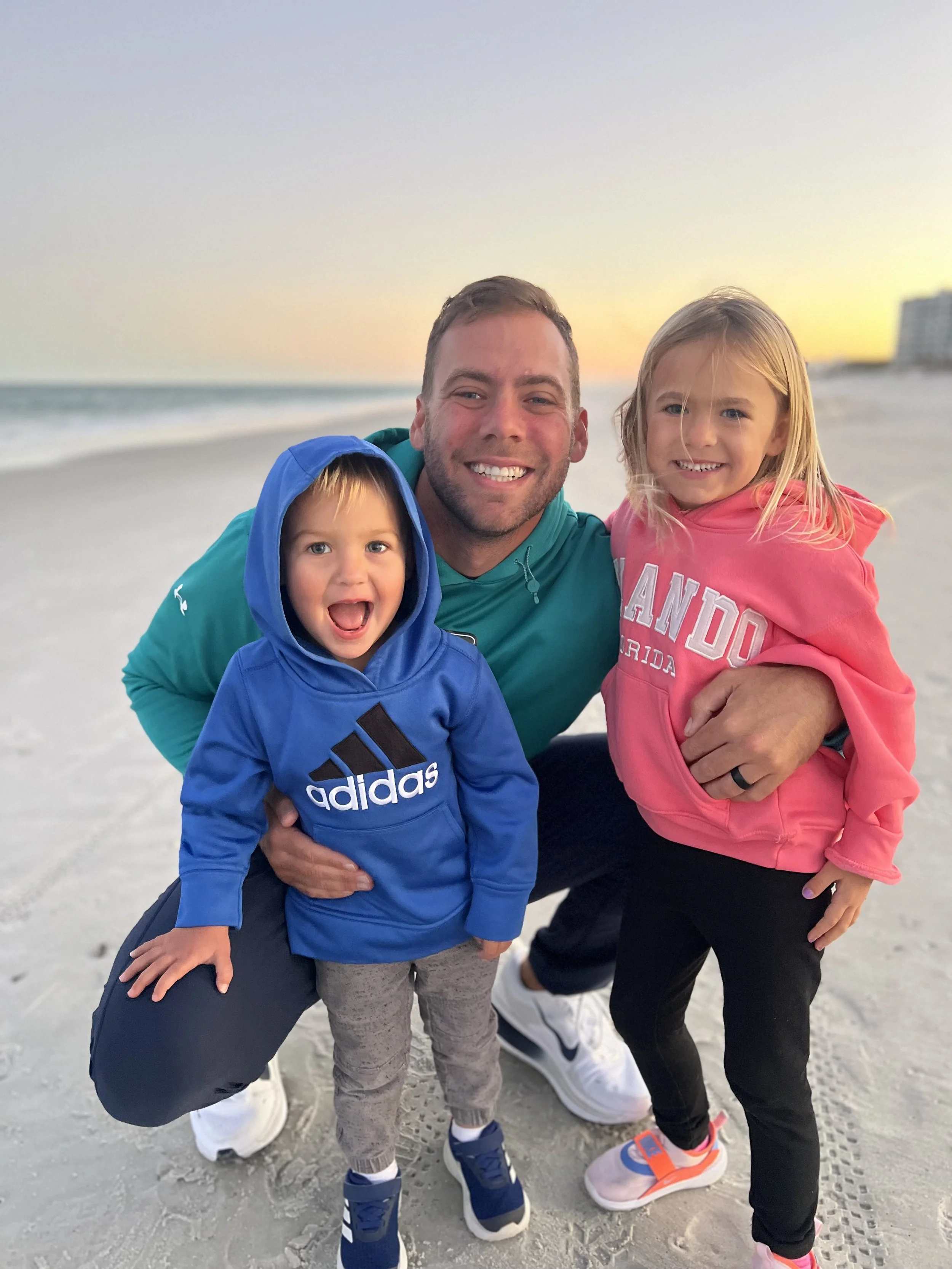 A man with two children smiling on the beach at sunset, one wearing a blue hoodie and the other in a pink hoodie.
