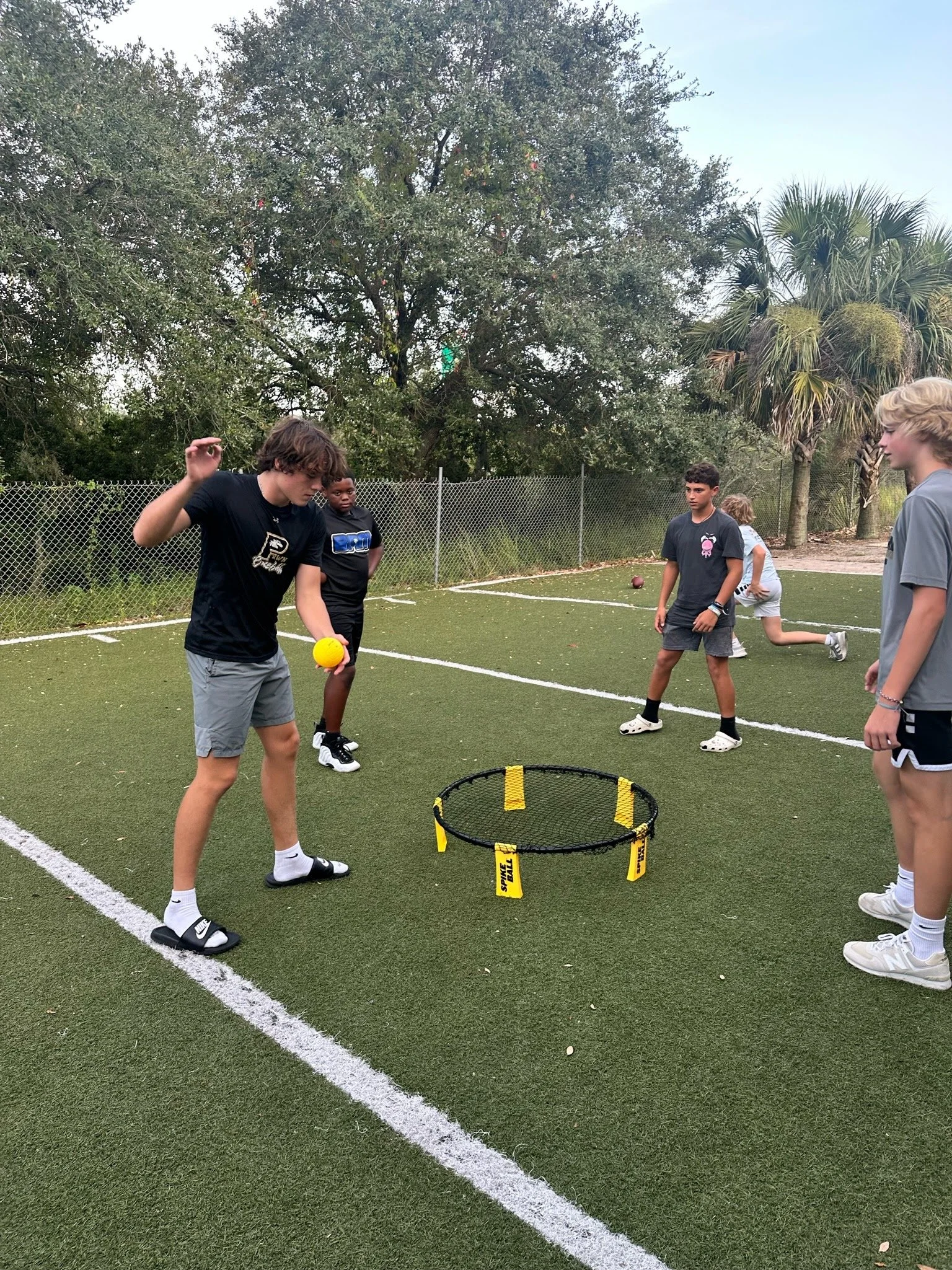 Group of children playing spikeball on an outdoor sports field surrounded by trees.