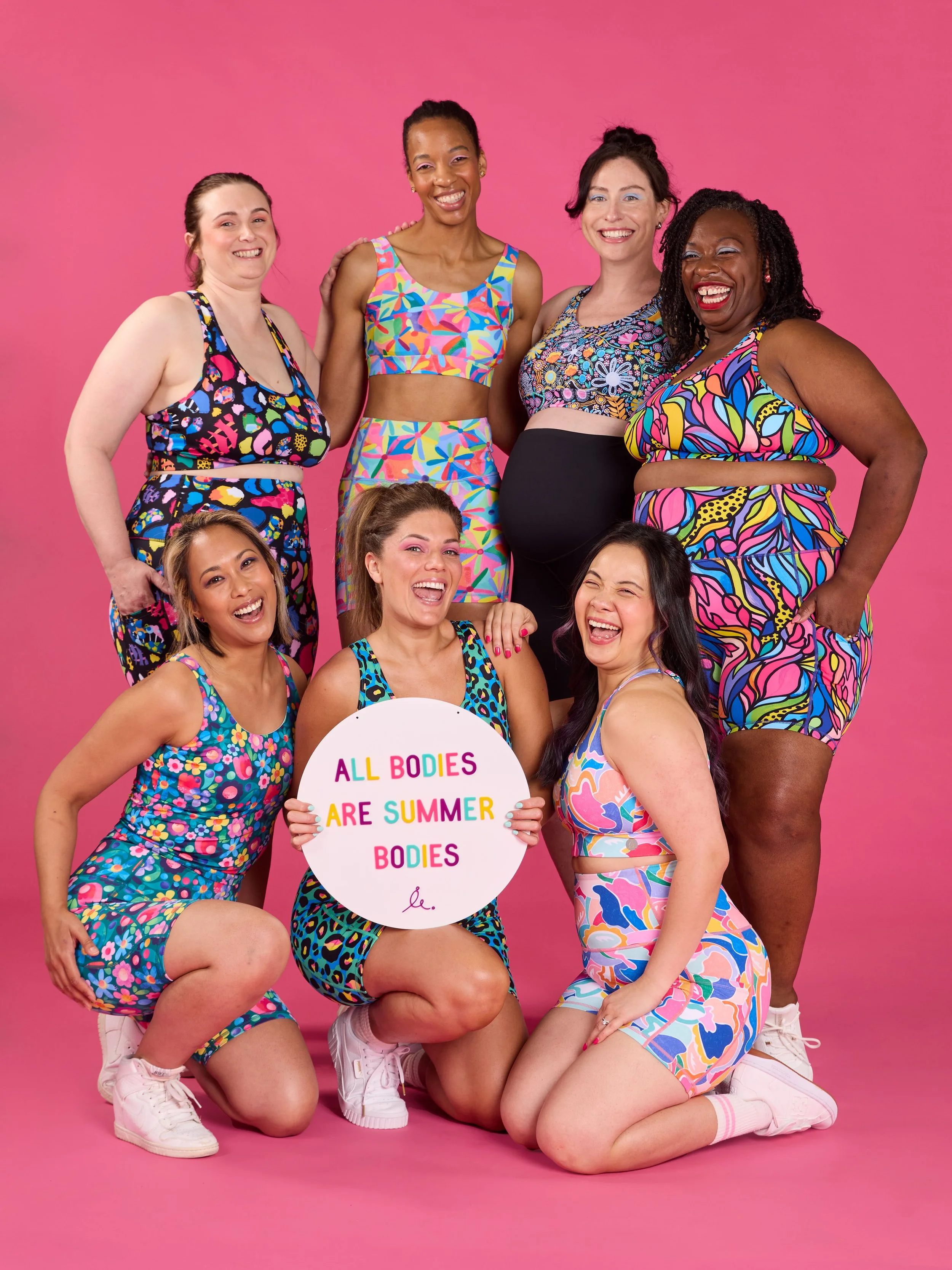A diverse group of nine women wearing colorful summer outfits, posing together against a pink background, with one woman holding a sign that reads, 'All bodies are summer bodies'.
