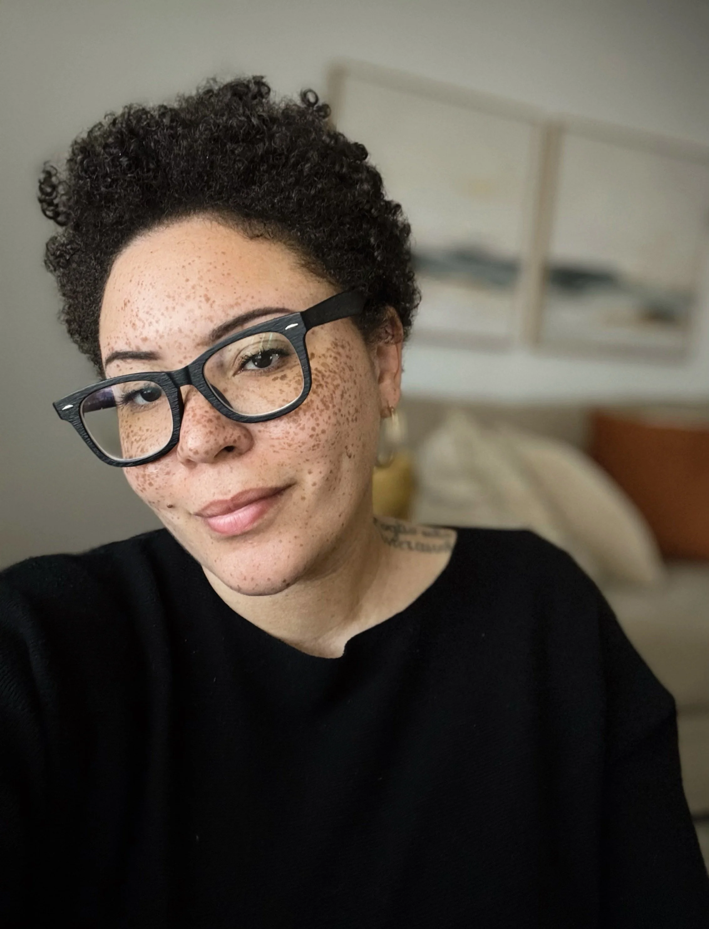 A woman with short curly hair, wearing black glasses and a black top, smiling in a softly lit room with a bed and artwork in the background.