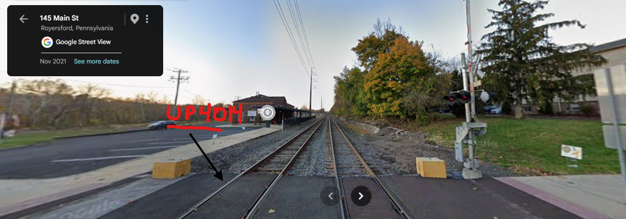 View looking down train tracks on Main Street in Royersford, Pennsylvania, with a railroad crossing signal and trees on the right side of the tracks.