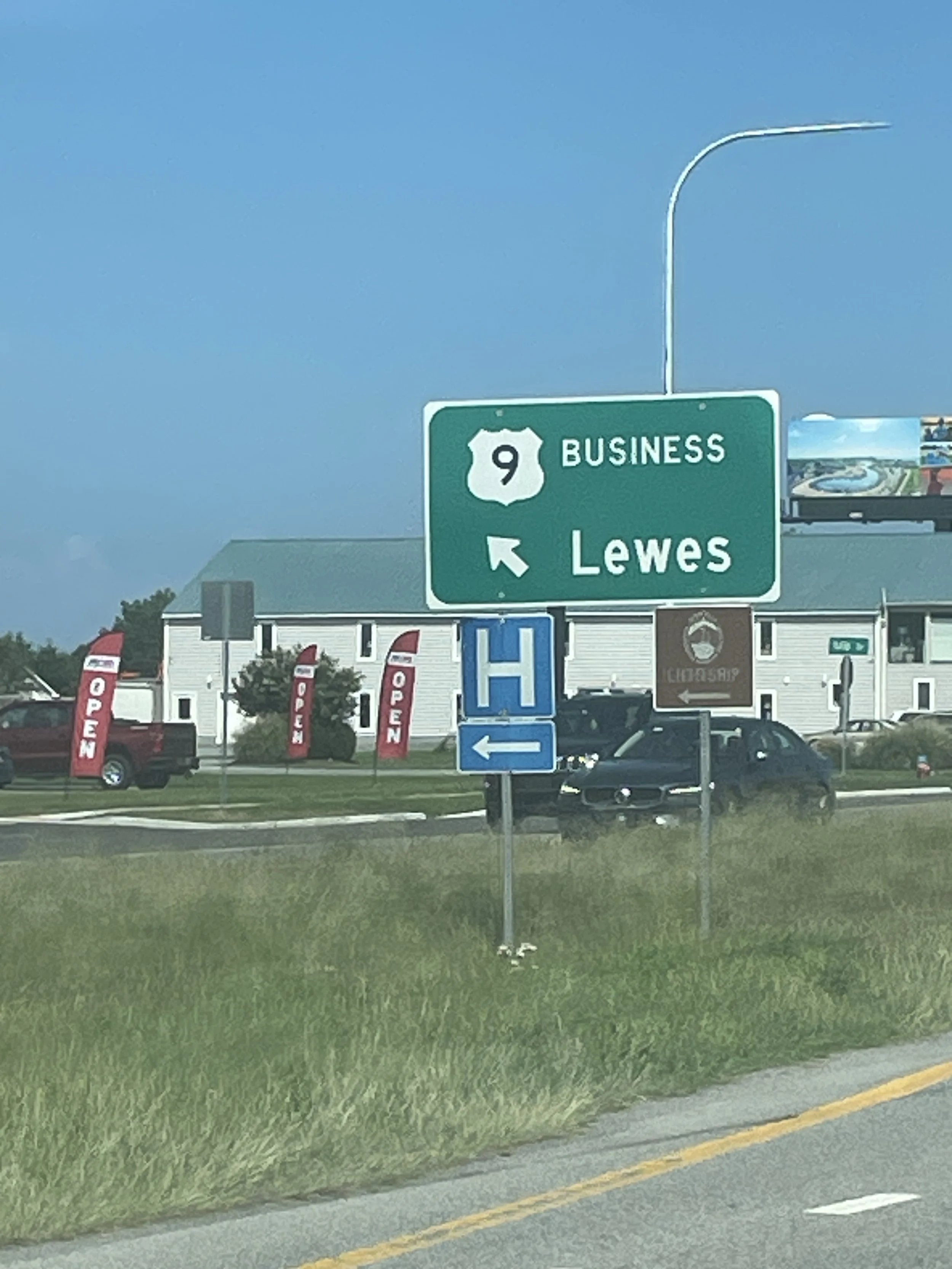 Road sign indicating US Highway 9 Business towards Lewes, with nearby buildings, parking lot, and cars, under a clear blue sky.