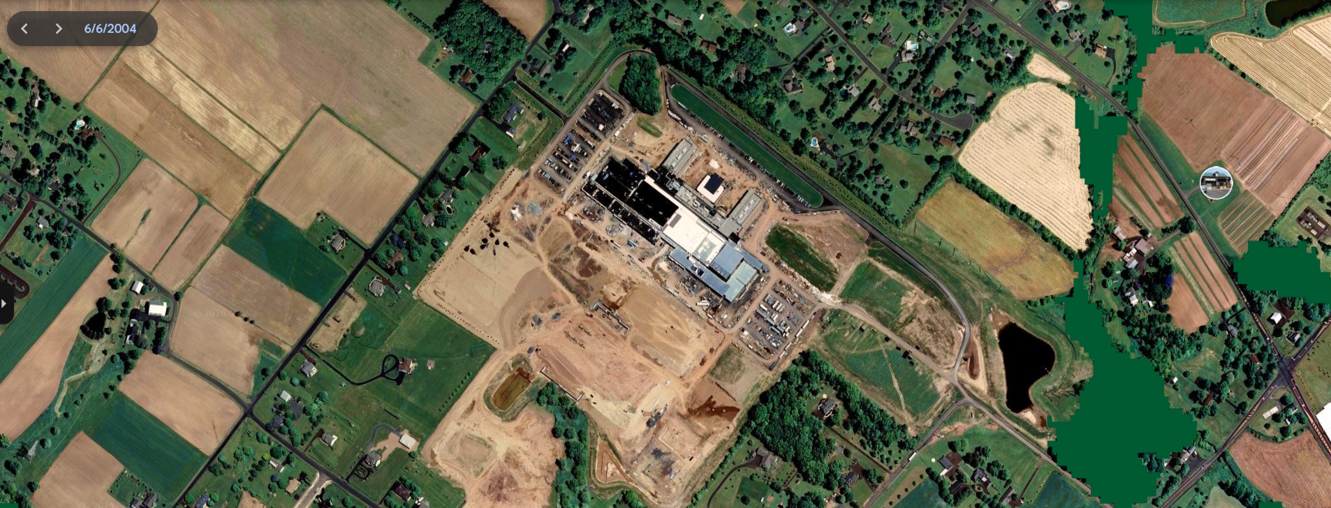 Aerial view of a construction site surrounded by farmland and residential neighborhoods with roads and trees.