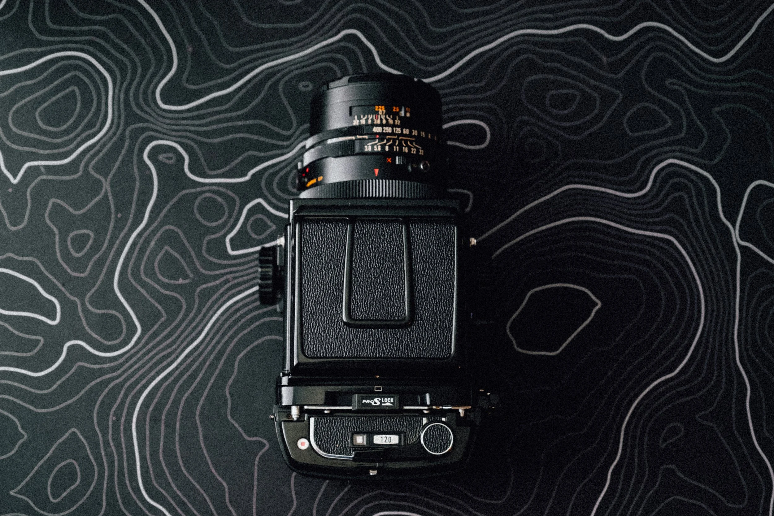 Top view of a vintage film camera with a black textured body and lens on a black surface with white contour topographic map lines.