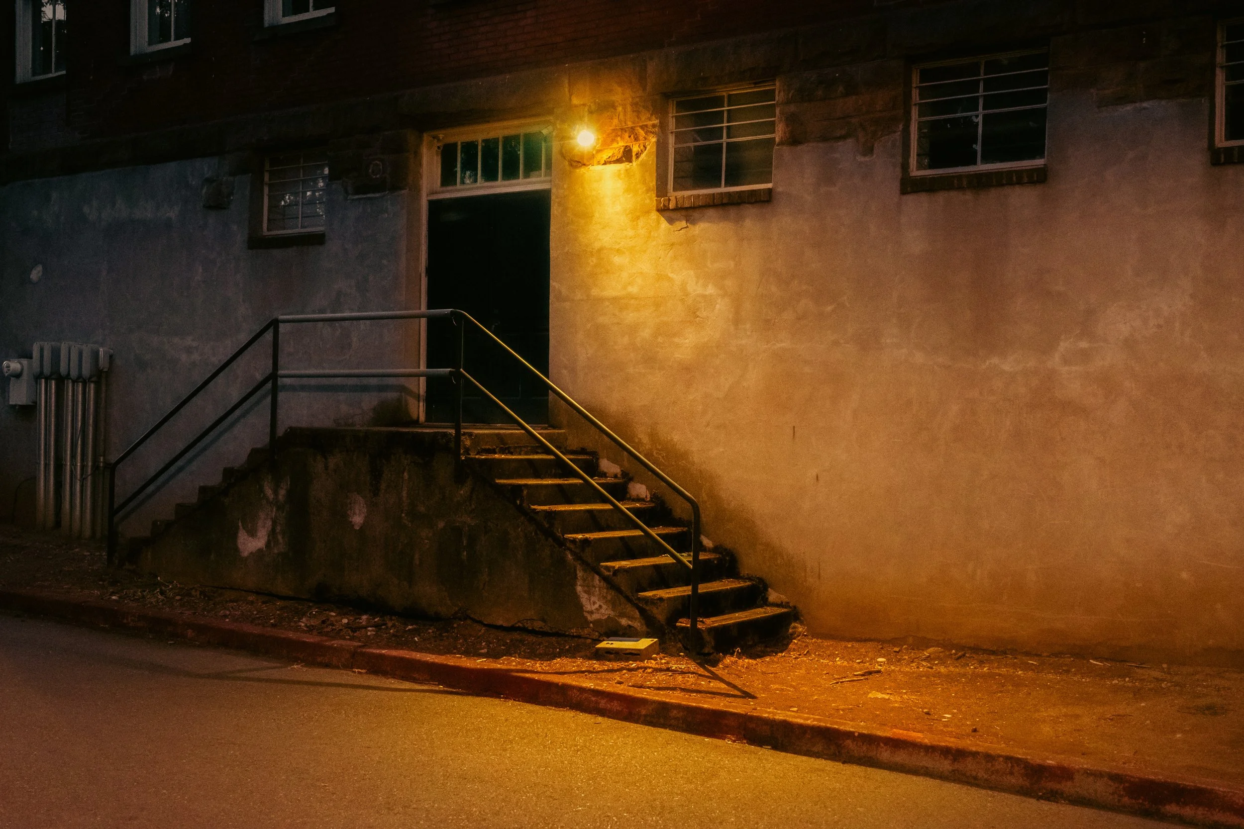 The Night By AJ Miranda Low-light, cinematic photograph of historic Vancouver, WA architecture during blue hour, using ambient light and deep chiaroscuro shadows to create an eerie film aesthetic.