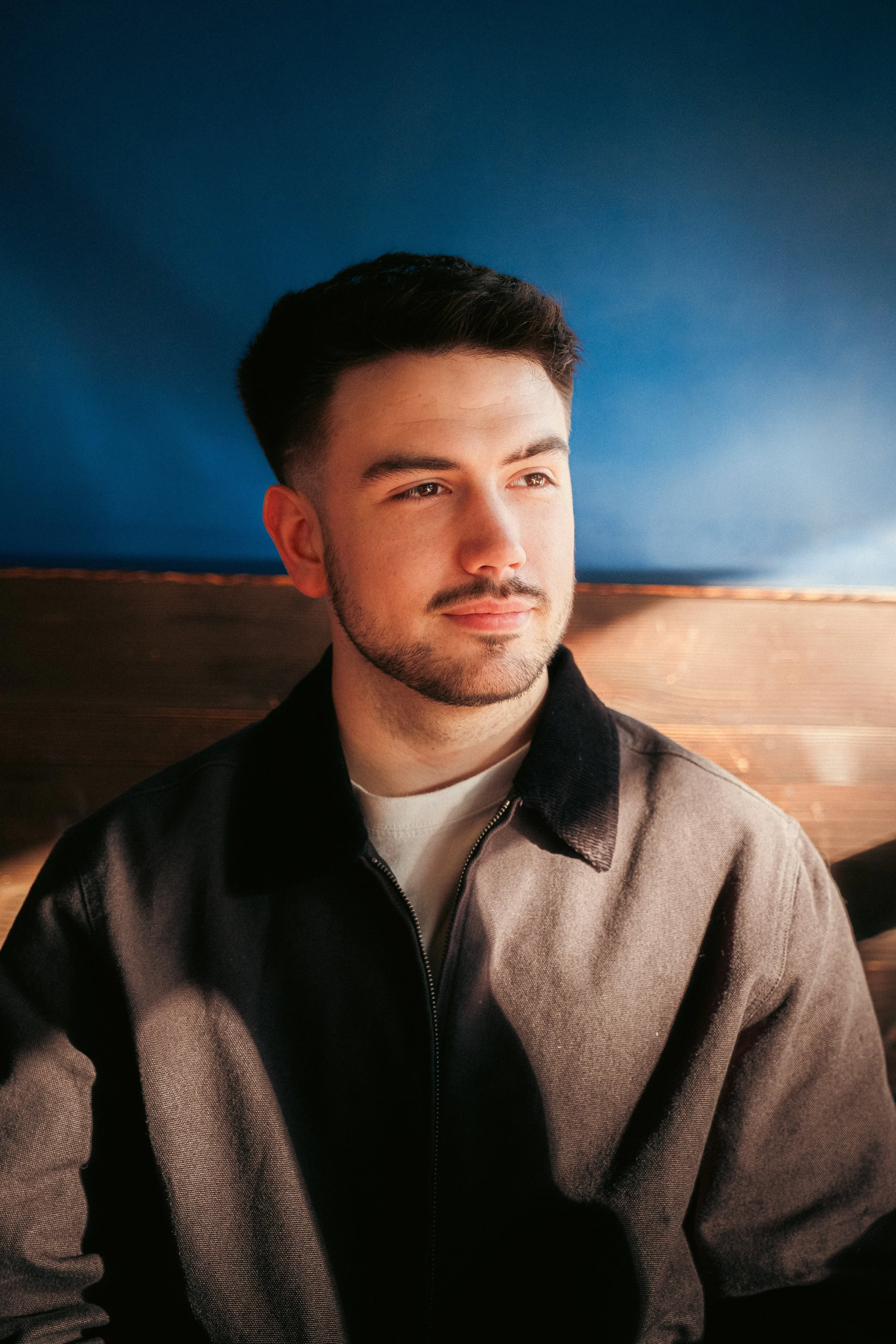 Portrait of a young man with dark hair, light beard, wearing a brown jacket, sitting indoors near a blue wall.
