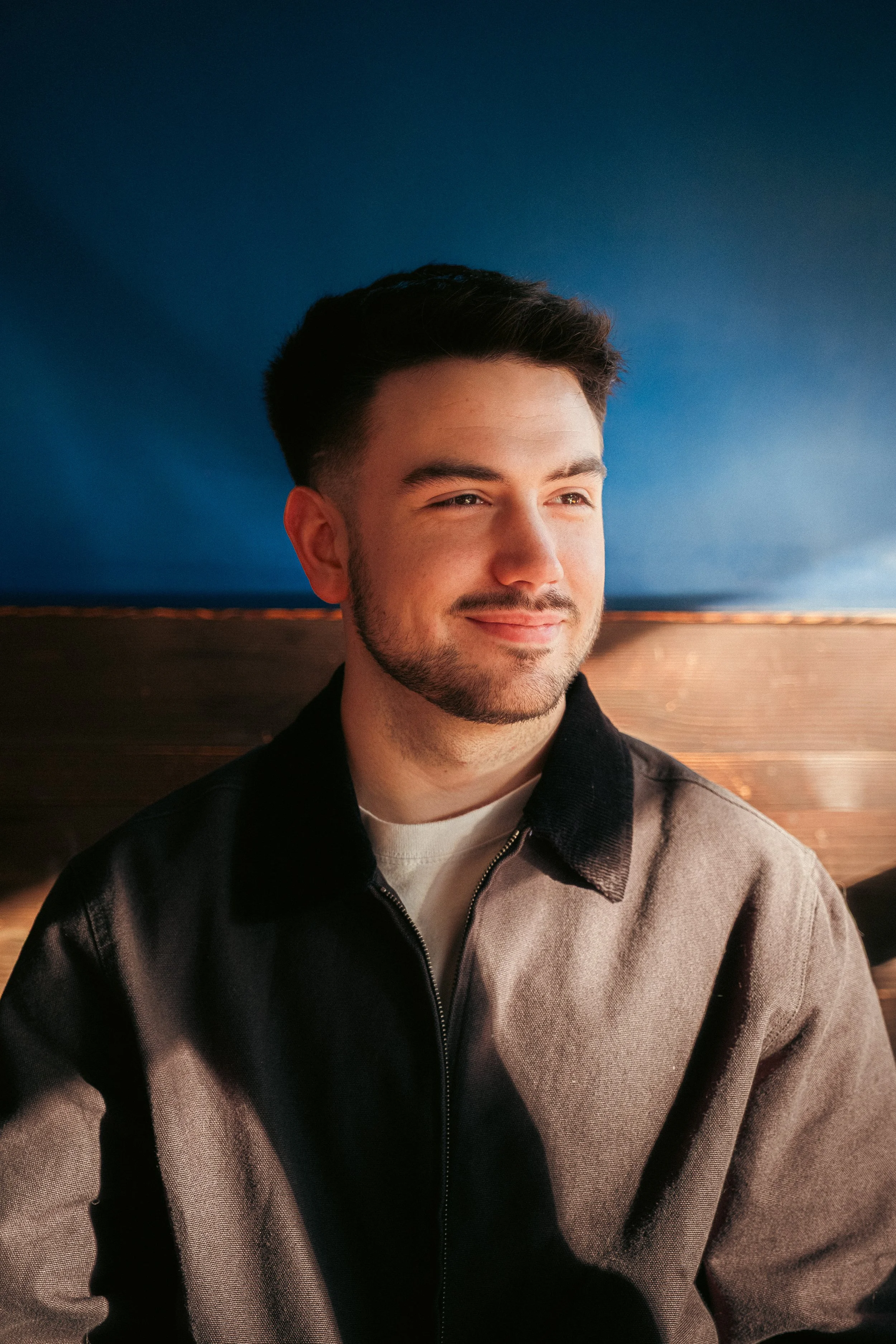 A young man with short dark hair, a beard, and a mustache, smiling and looking to the side. He is wearing a black and brown jacket with a zipper, against a background featuring a wooden surface and a blue wall.