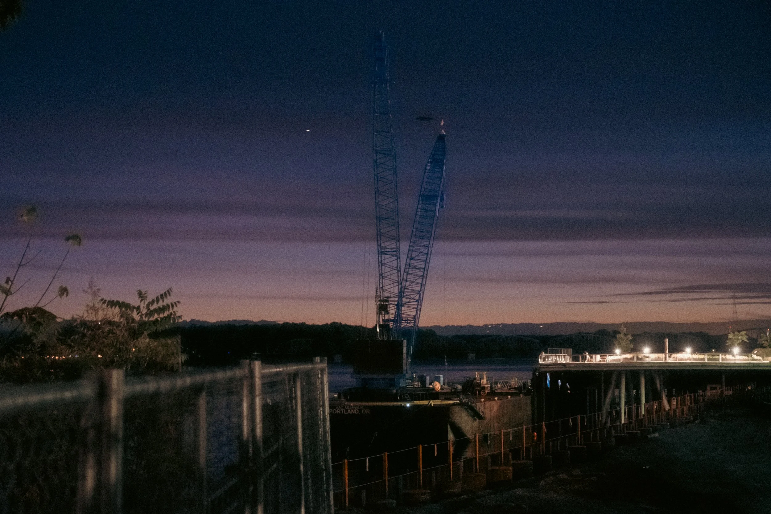 Cinematic, low-light photograph from "Sunset Blues" series by AJ Miranda, featuring a bloomy moon and silhouetted trees against a pink and orange Golden Hour skyscape.