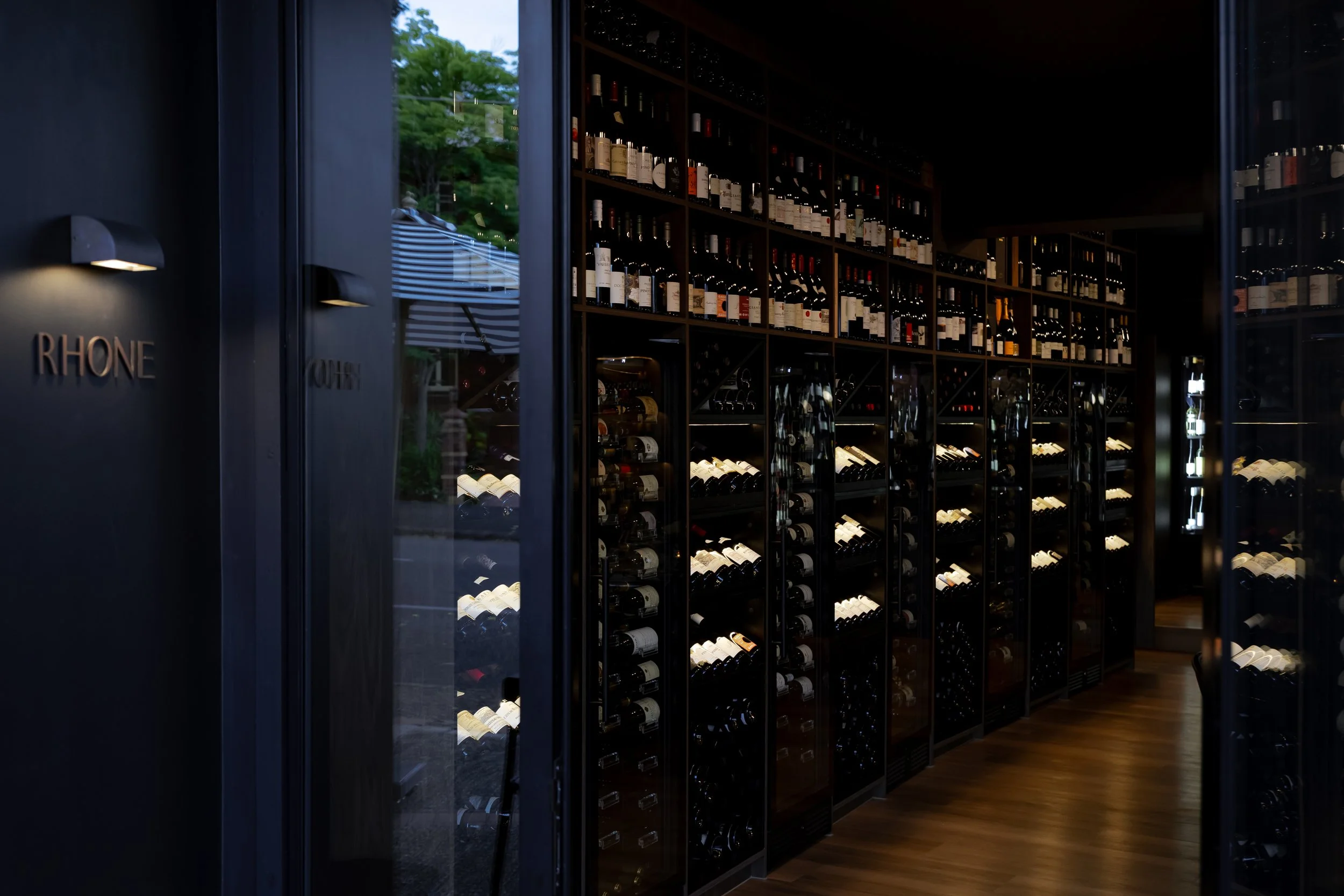 Wine cellar with floor-to-ceiling racks and fridges filled with bottles of wine, visible through a glass door with the word 'RHONE' on the wall next to it.