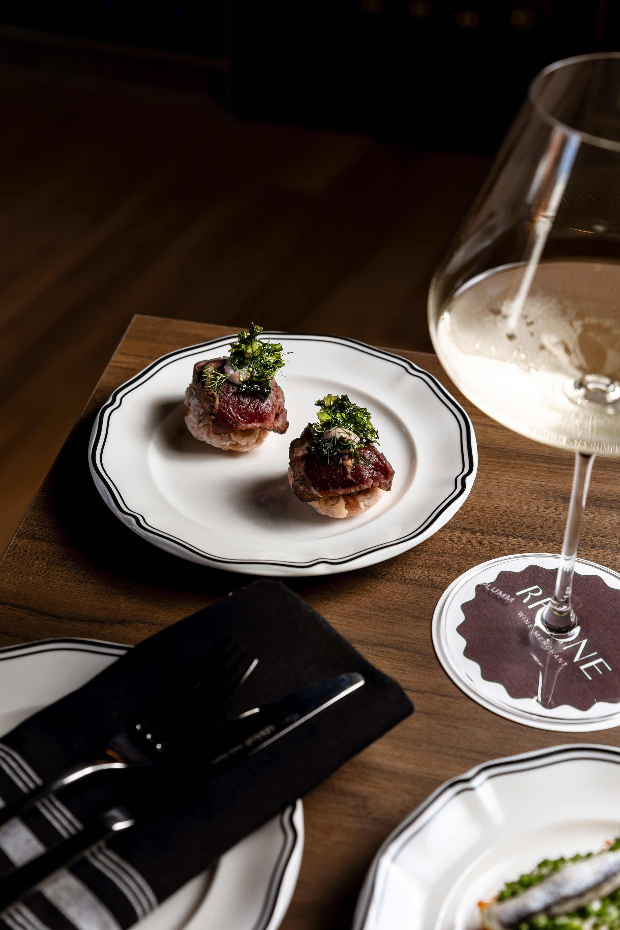 Two pieces of beef carpaccio topped with greens on a white plate, a glass of white wine on a wooden table, black napkin with utensils, and additional plates.