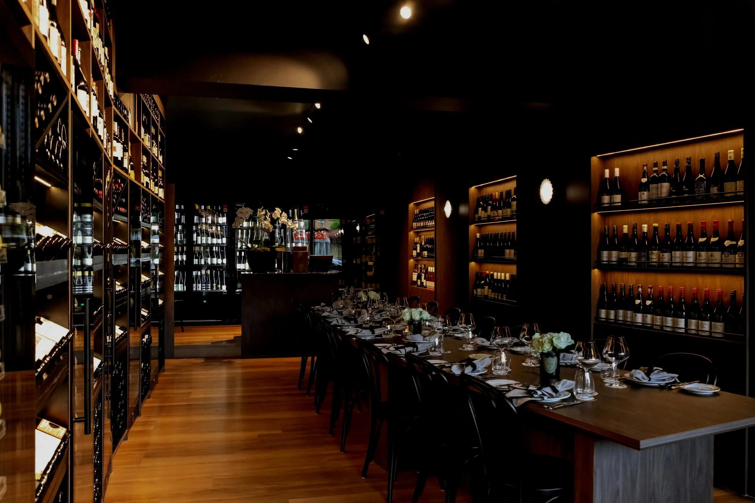 An elegant dining room with a long wooden table set with wine glasses, plates, napkins, and floral centerpieces, surrounded by black chairs. Shelves filled with wine bottles line the dark walls.