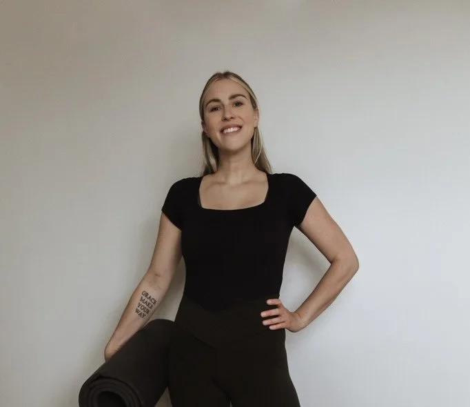 A woman with long brown wavy hair smiling, wearing a grey Nike sweatshirt with 'Pilates Boutique Bowmanville' text, standing in front of a beige wall.