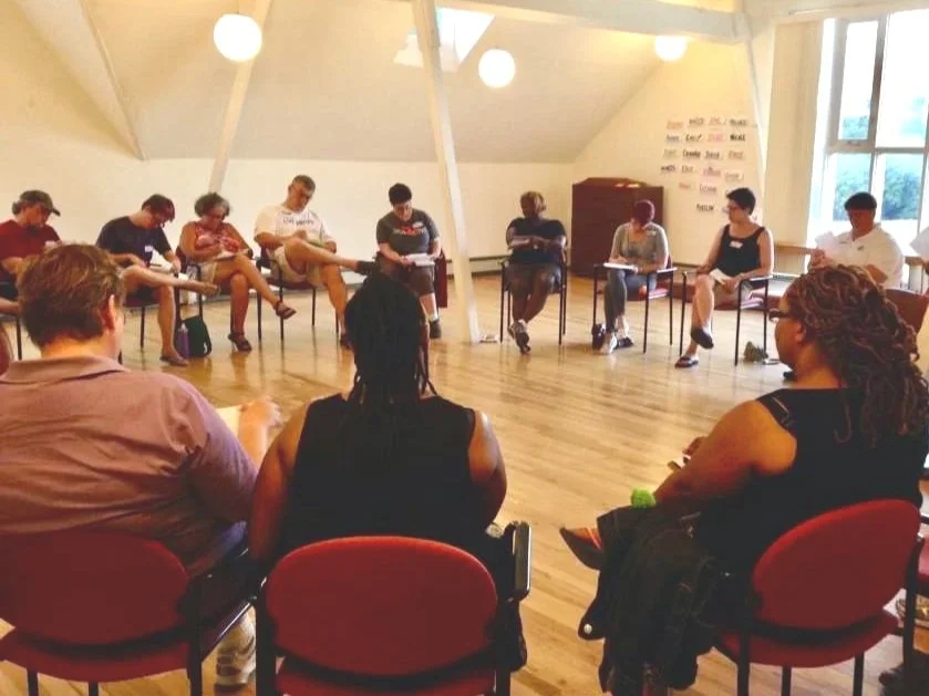 Group of people sitting in a circle in a room with wooden floor and large windows, engaged in a discussion or meeting.