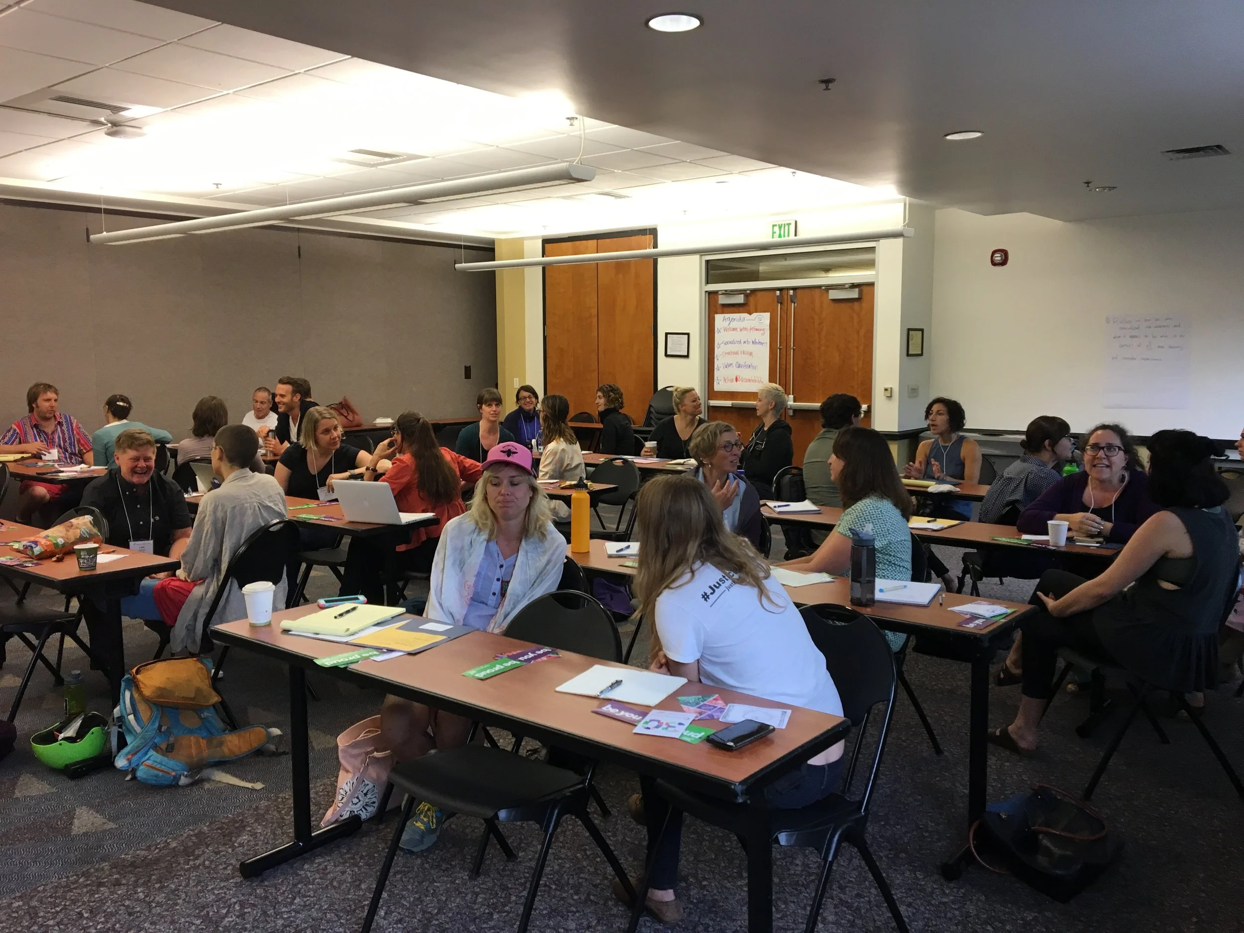 Evangeline Weiss Consulting | A group of people sitting at tables in a conference room or classroom, engaging in conversations and working on laptops and papers.