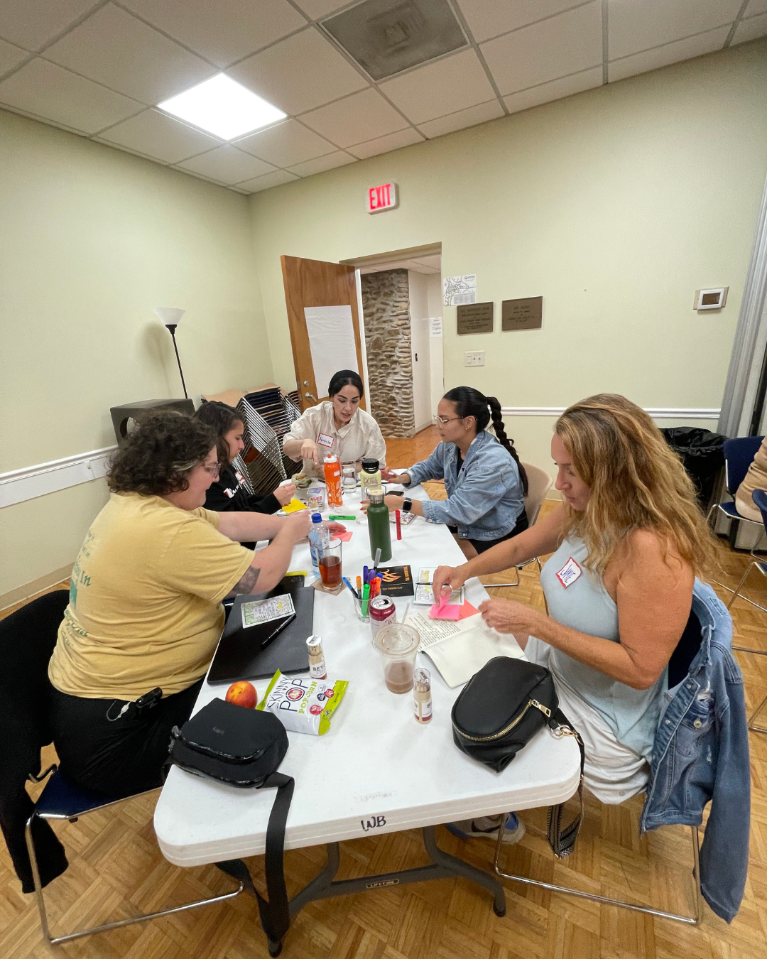 Evangeline Weiss Consulting | Group of five women sitting around a table engaging in arts and crafts activities, with various supplies like markers, paper, water bottles, and snacks on the table, in a room with pale yellow walls and wooden flooring.