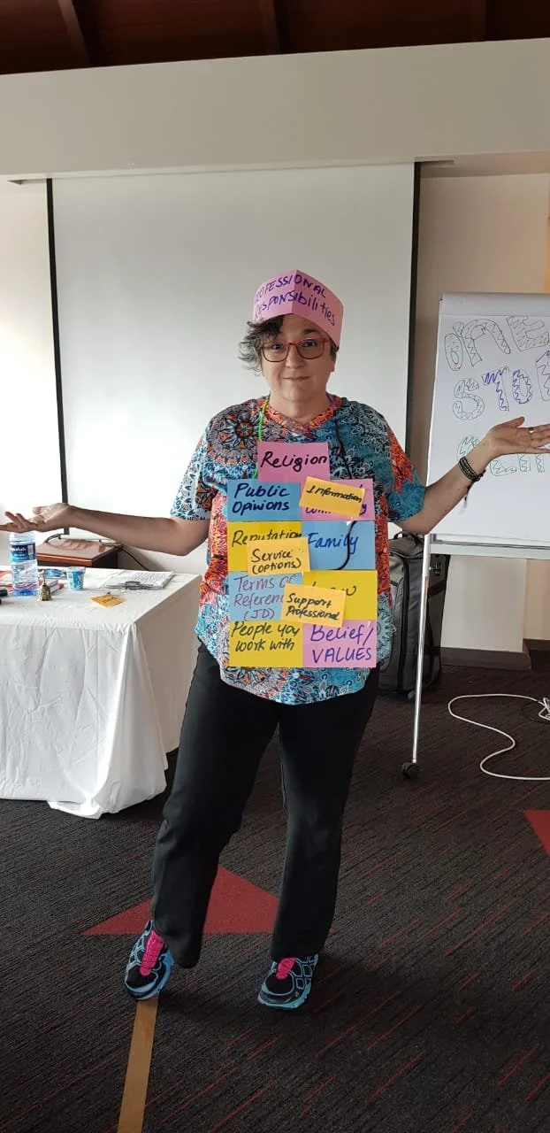Evangeline Weiss Consulting | A woman standing in a room with a whiteboard in the background, wearing a pink hat that says 'Professional Responsibilities'.