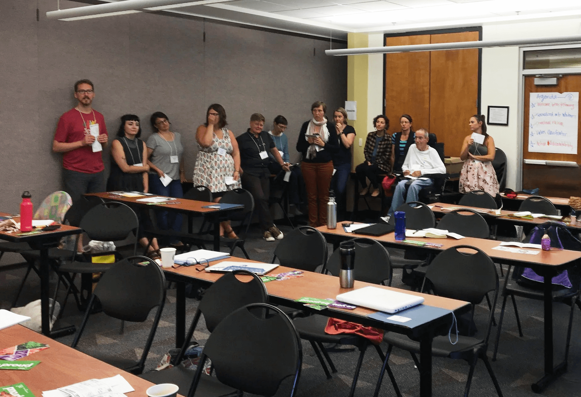 Evangeline Weiss Consulting | Group of adults standing in a conference room, some sitting, with tables, chairs, and a whiteboard with notes in the background.
