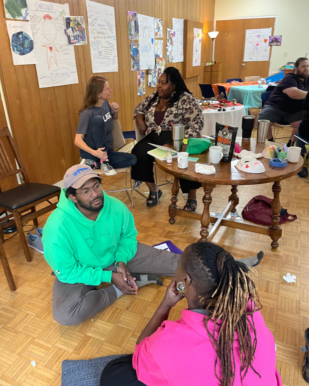 Evangeline Weiss Consulting | People engaging in conversations in a room decorated with posters and artwork on the walls, with a large wooden table cluttered with cups, papers, and personal items, and others sitting and talking in the background.