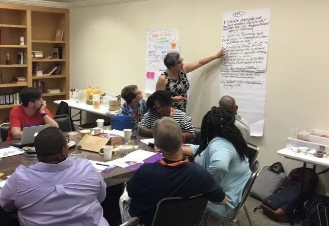 Evangeline Weiss Consulting | A woman standing at the front of a room pointing at a large sheet of paper on the wall, while six people seated around a table listen and take notes during a meeting or workshop.