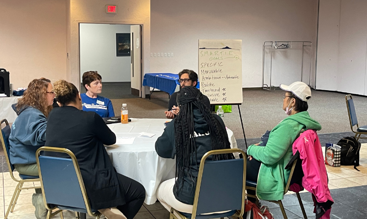 Evangeline Weiss Consulting | Five people sitting around a round table in a meeting room, with a whiteboard and a small refreshment table in the background.