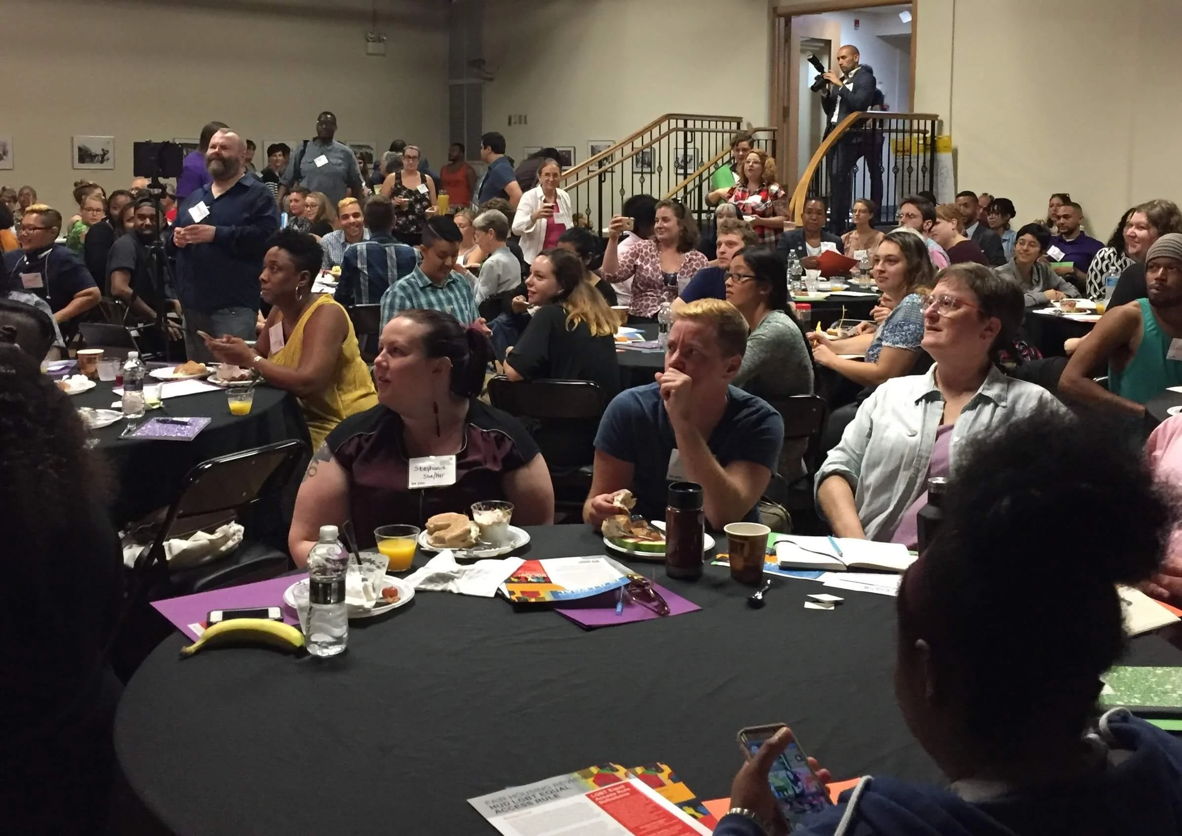 Evangeline Weiss Consulting | A large group of people in a conference room, some seated at tables with food and drinks, others standing or taking photos. A man is on a staircase with a camera, and the room is filled with diverse attendees.