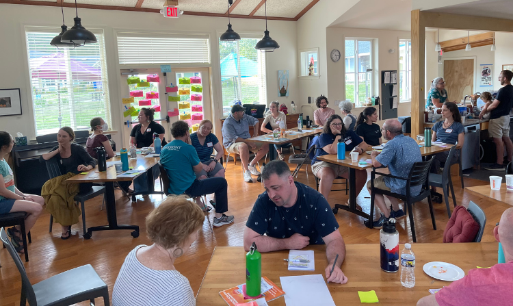 Evangeline Weiss Consulting | People sitting at tables in a brightly lit room, engaged in conversation, with some holding drinks or snacks, and colorful sticky notes on a window in the background.