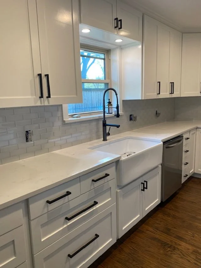 Modern white kitchen with black handles, farmhouse sink, marble countertops, and a window above the sink showing trees outside.
