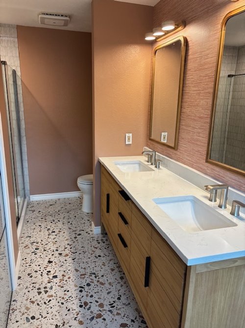 Modern bathroom with double vanity sinks, wooden cabinet, two mirrors, beige walls, terrazzo flooring, toilet, and shower stall.