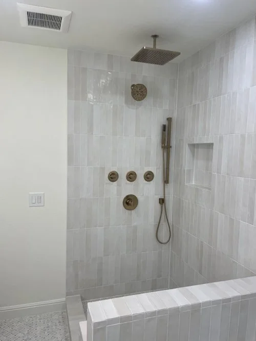 Modern walk-in shower with a ceiling-mounted rain showerhead, a handheld shower with a sliding bar, and wall controls, surrounded by light-colored tiles and a small built-in niche.