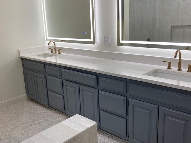 Modern bathroom vanity with blue cabinets, two white sinks, and gold fixtures. Large mirrors are above the sinks, and there is a small white ottoman in front.