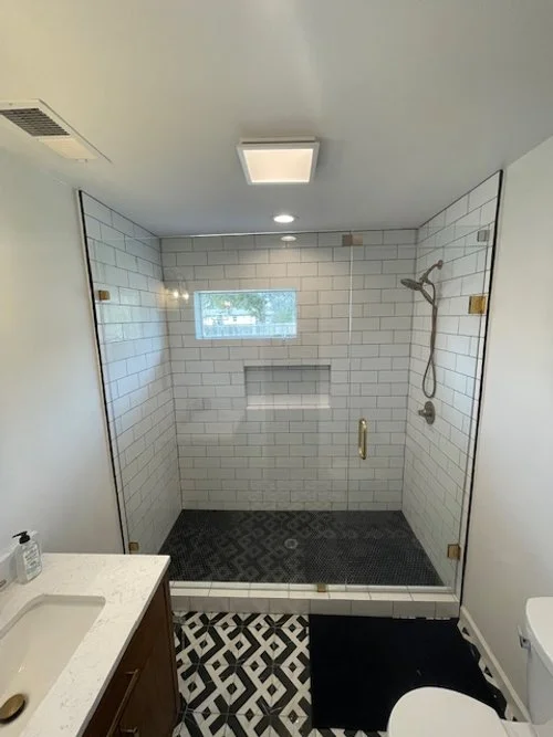 Glass-enclosed walk-in shower with white subway tiles, a small window, a wall-mounted showerhead, and built-in shelves, in a bathroom with black and white patterned floor tiles.