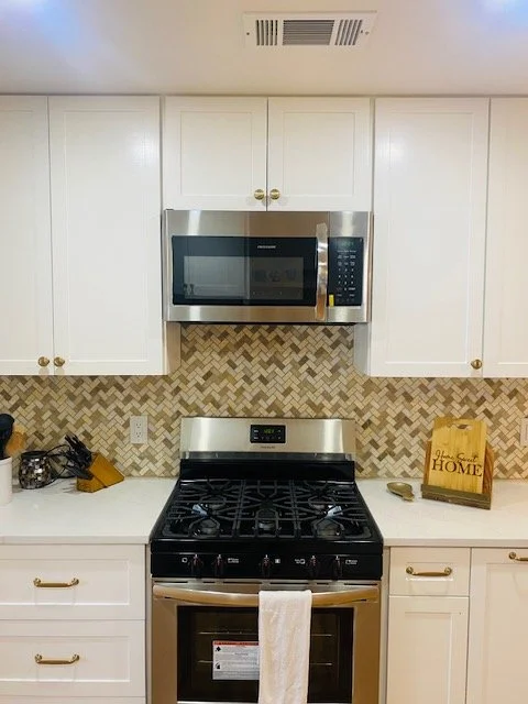 Kitchen with white cabinets, stainless steel microwave and gas stove, beige and brown herringbone backsplash, and various small kitchen items on the countertop.