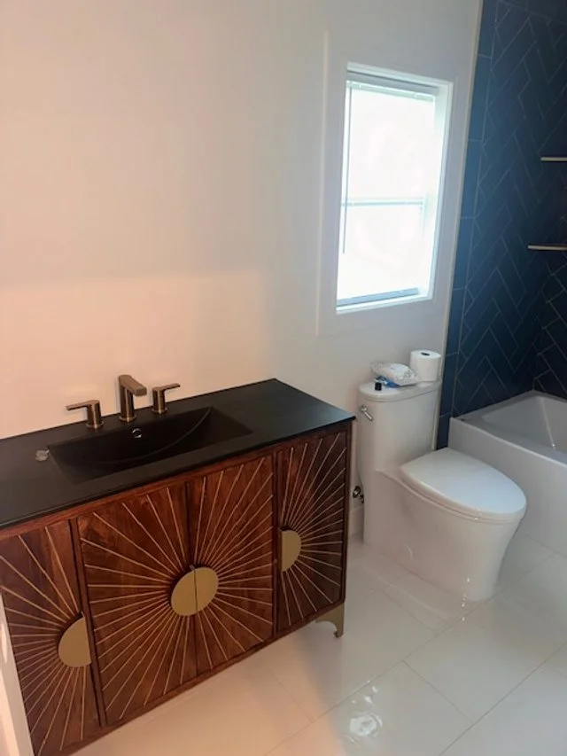 Modern bathroom with a dark wood vanity, black sink, and gold accents, along with a white toilet and a bathtub, illuminated by natural light from a window.