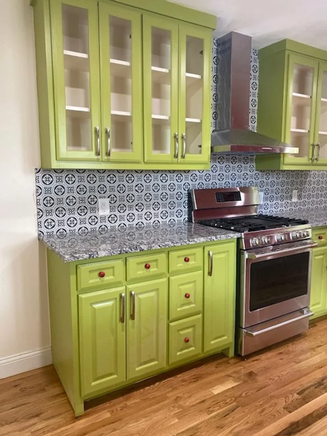 Green kitchen cabinets with glass-paneled upper doors and red knobs, a granite countertop, a stainless steel stove, a decorative black and white tiled backsplash, and hardwood flooring.
