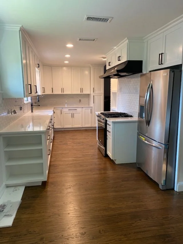 Empty white kitchen with wooden flooring, stainless steel refrigerator, stove, and white cabinets, with a countertop and under-cabinet lighting.