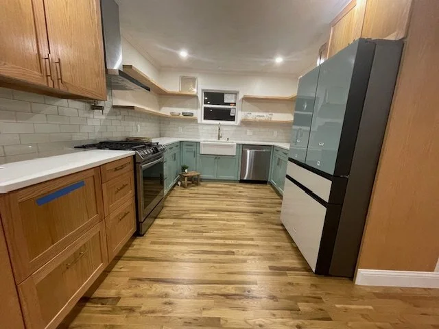 A modern kitchen with wooden and teal cabinets, stainless steel appliances, and wood flooring.